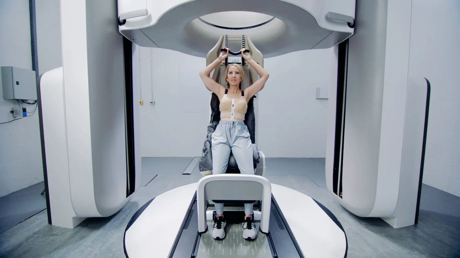 Patientin sitzt aufrecht im Leo-Cancer-Care-System, die Arme über dem Kopf gelagert, darüber der Ring des Upright-CT